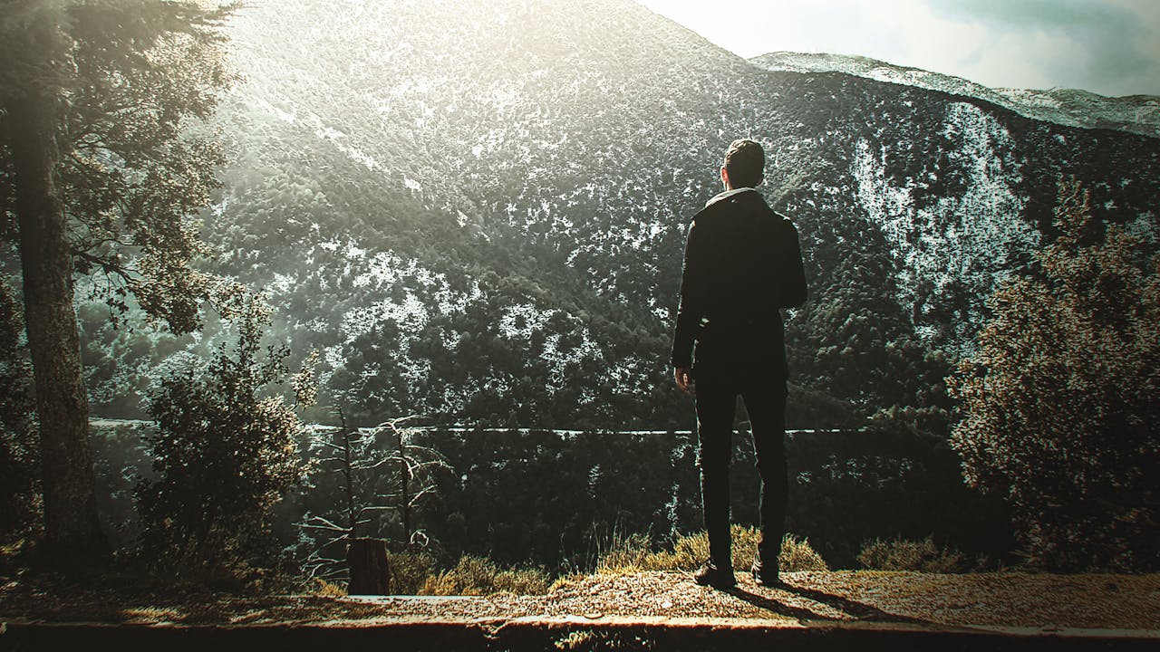 A person stands alone, observing a sunlit mountainous landscape during the day.
